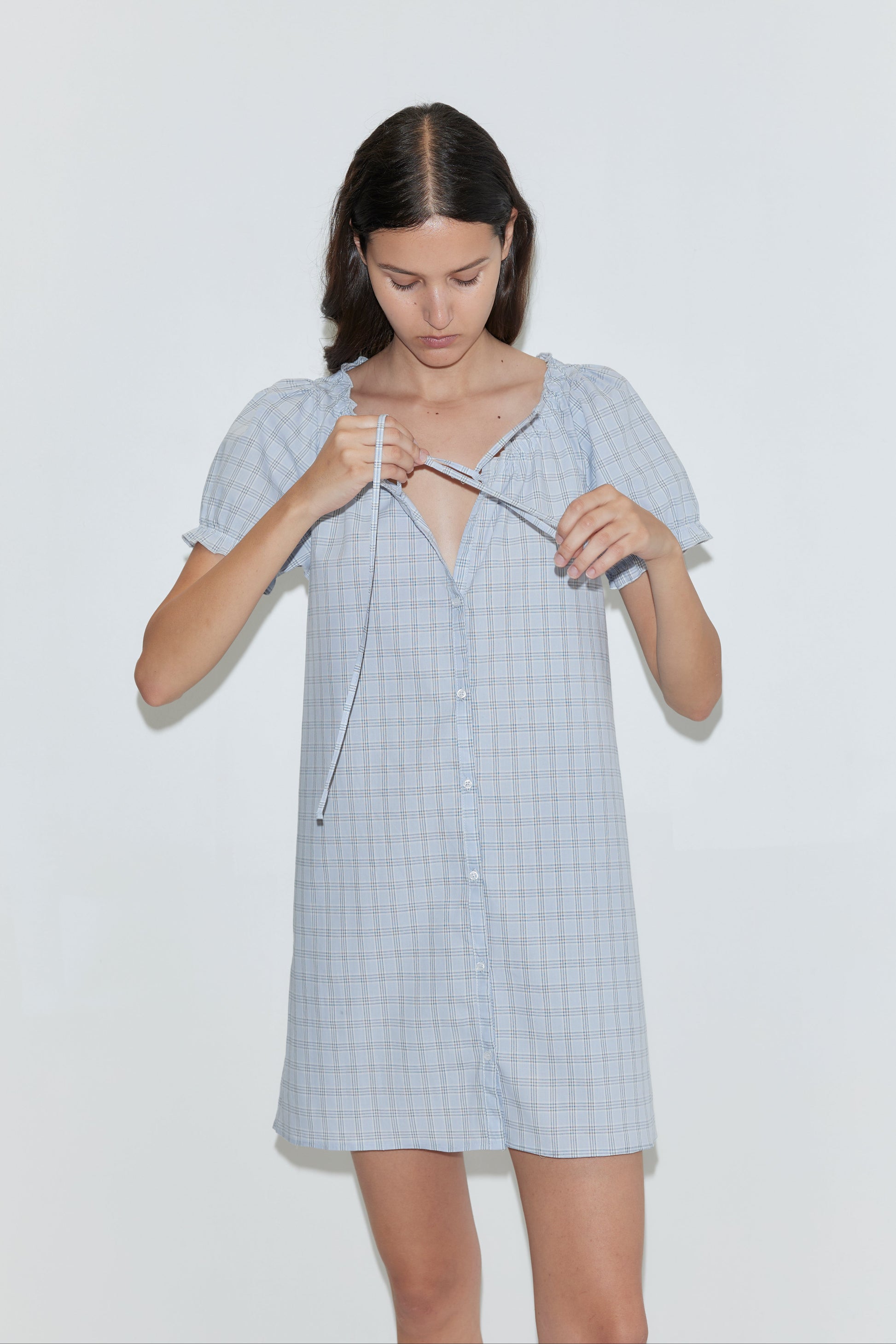 A woman with straight brown hair wears the Buttoned Mini Dress - Powder Check from Deiji Studios, tying the front neckline as she stands against a plain white background.