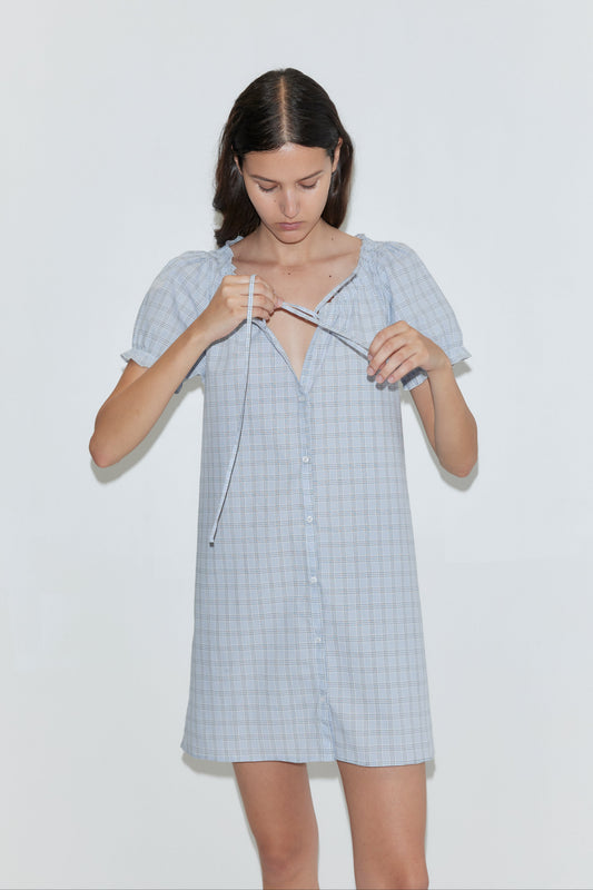 A woman with straight brown hair wears the Buttoned Mini Dress - Powder Check from Deiji Studios, tying the front neckline as she stands against a plain white background.