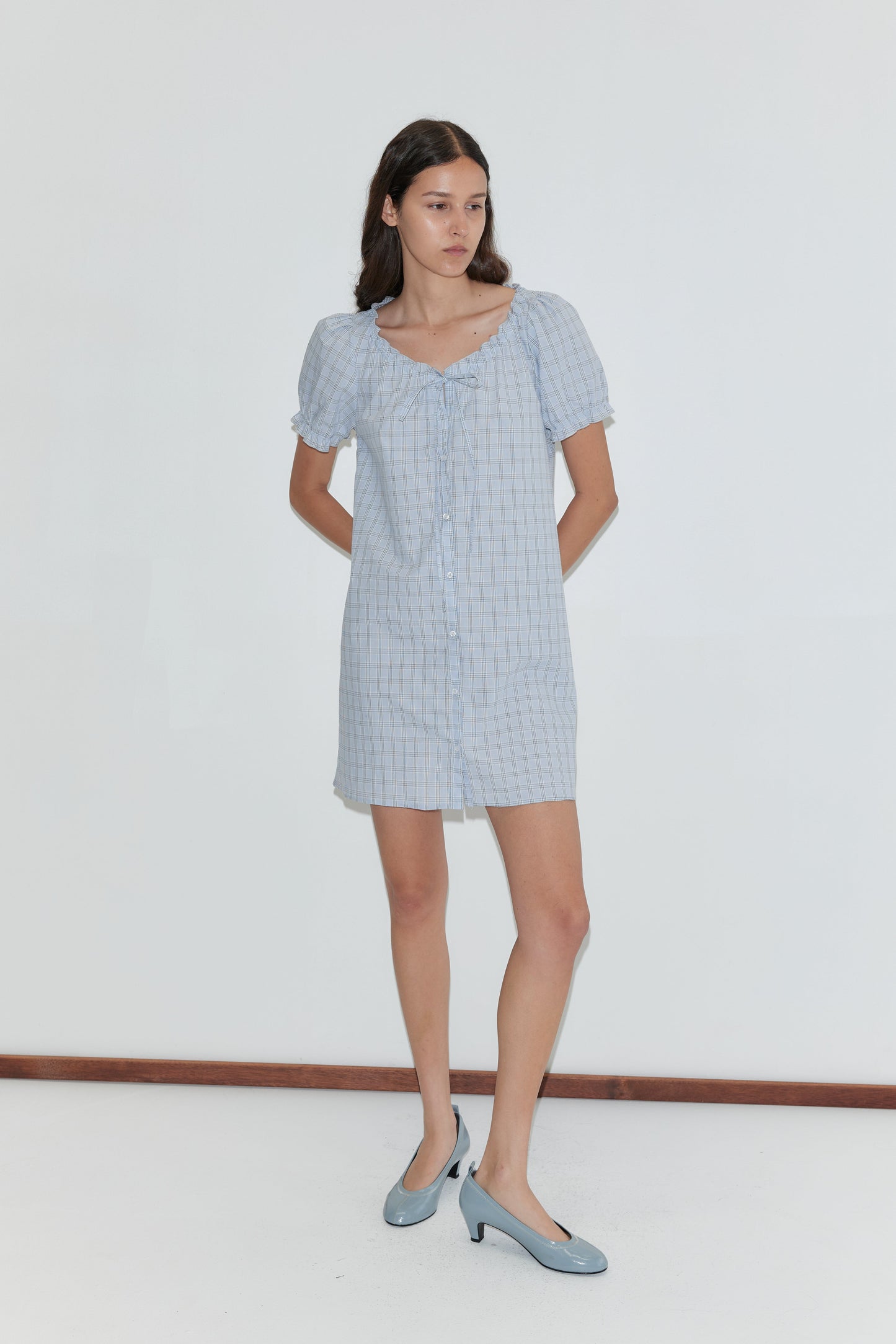 A woman wears the Buttoned Mini Dress - Powder Check by Deiji Studios, featuring a tie neckline. She pairs it with light blue heels and poses against a plain white background.