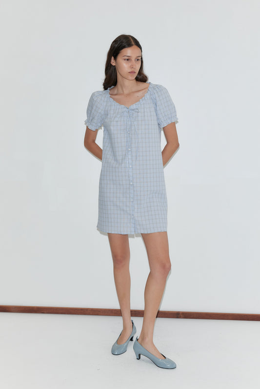 A woman wears the Buttoned Mini Dress - Powder Check by Deiji Studios, featuring a tie neckline. She pairs it with light blue heels and poses against a plain white background.