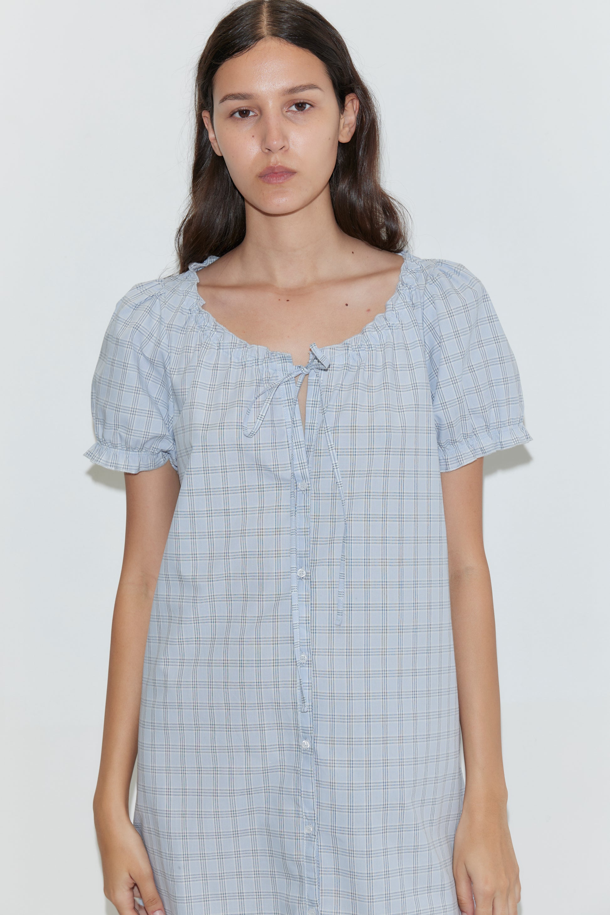 A woman with long, dark hair wears the Buttoned Mini Dress - Powder Check by Deiji Studios, featuring a light blue check pattern, scoop neckline, tie detail, and button-down front against a plain light background.