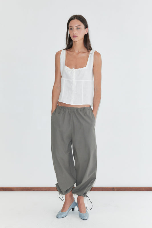 A woman with long dark hair stands against a plain white background wearing the Relax Tie Top - White by Deiji Studios, a white sleeveless linen top with a gathered drawstring neckline, with grey check trousers and pale blue heels.
