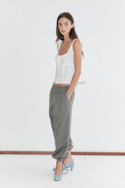 A woman with long dark hair stands in profile against a plain white background wearing the Relax Tie Top - White by Deiji Studios, a white sleeveless linen top, with grey check trousers and pale blue heels.