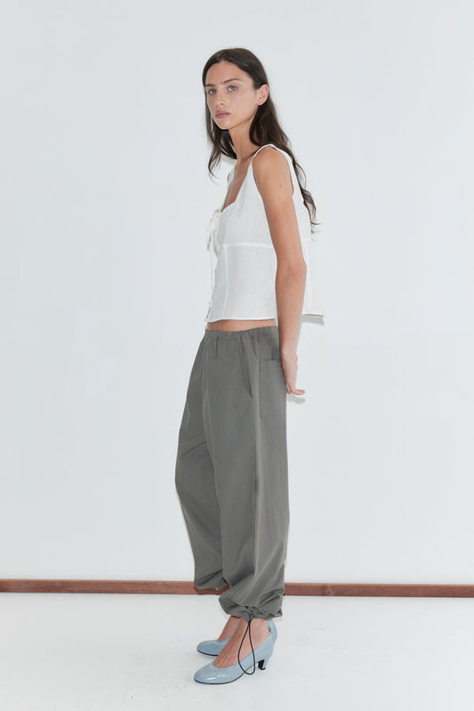 A woman with long dark hair stands in profile against a plain white background wearing the Relax Tie Top - White by Deiji Studios, a white sleeveless linen top with a drawstring tie neckline, with grey check trousers and pale blue heels.