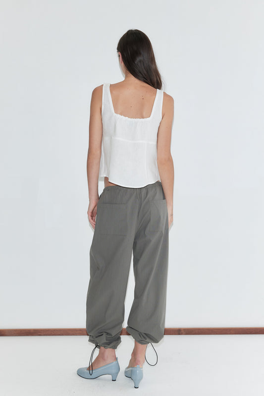 A woman with long dark hair stands with her back to the camera against a plain white background wearing the Relax Tie Top - White by Deiji Studios, a white sleeveless linen top, with grey check trousers and pale blue heels.