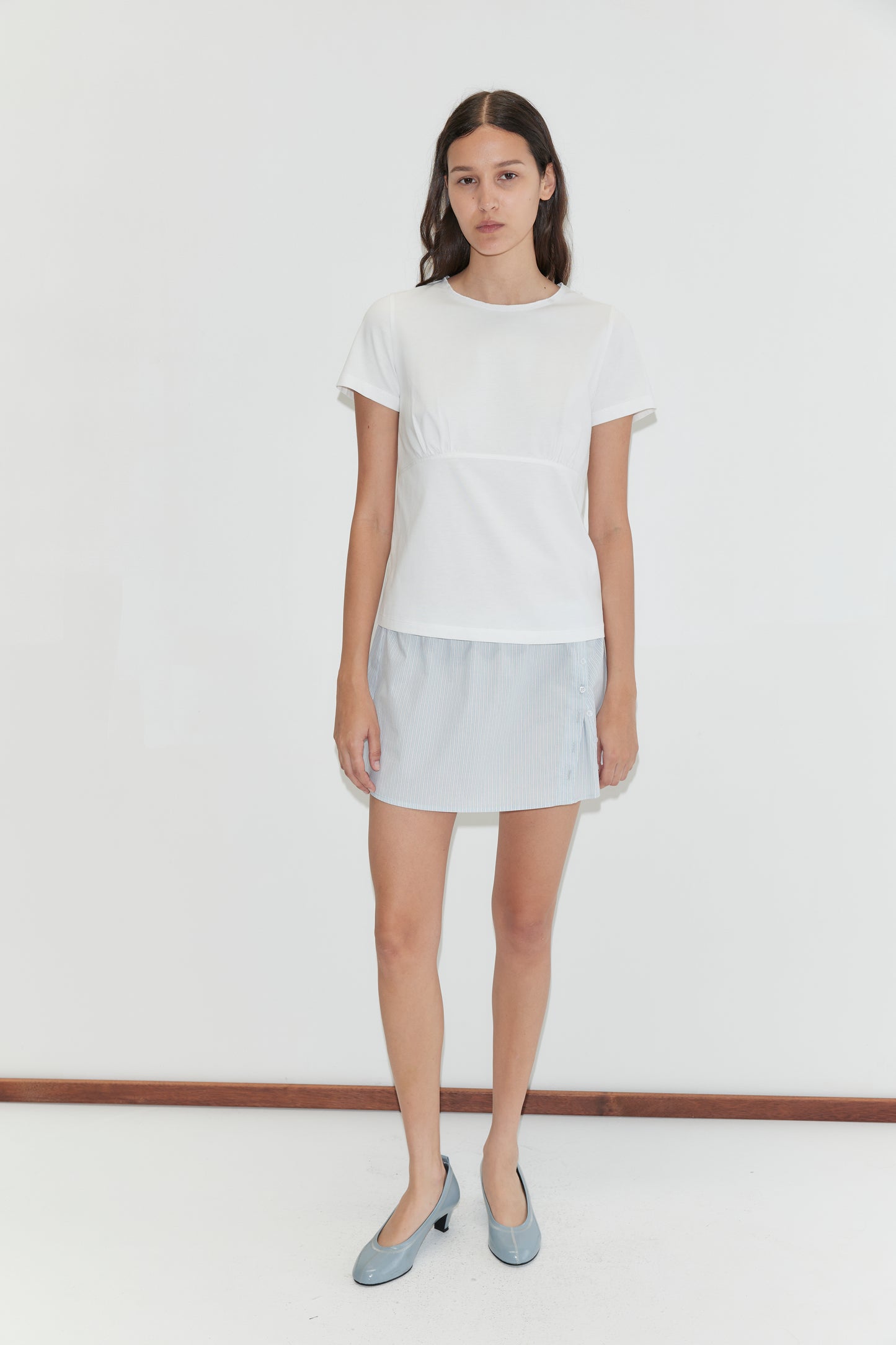 A woman with long dark hair and a neutral expression stands against a plain white background wearing the Pleat Split Skirt - Mist Stripe by Deiji Studios, a short-sleeved white top, and light blue flat shoes.