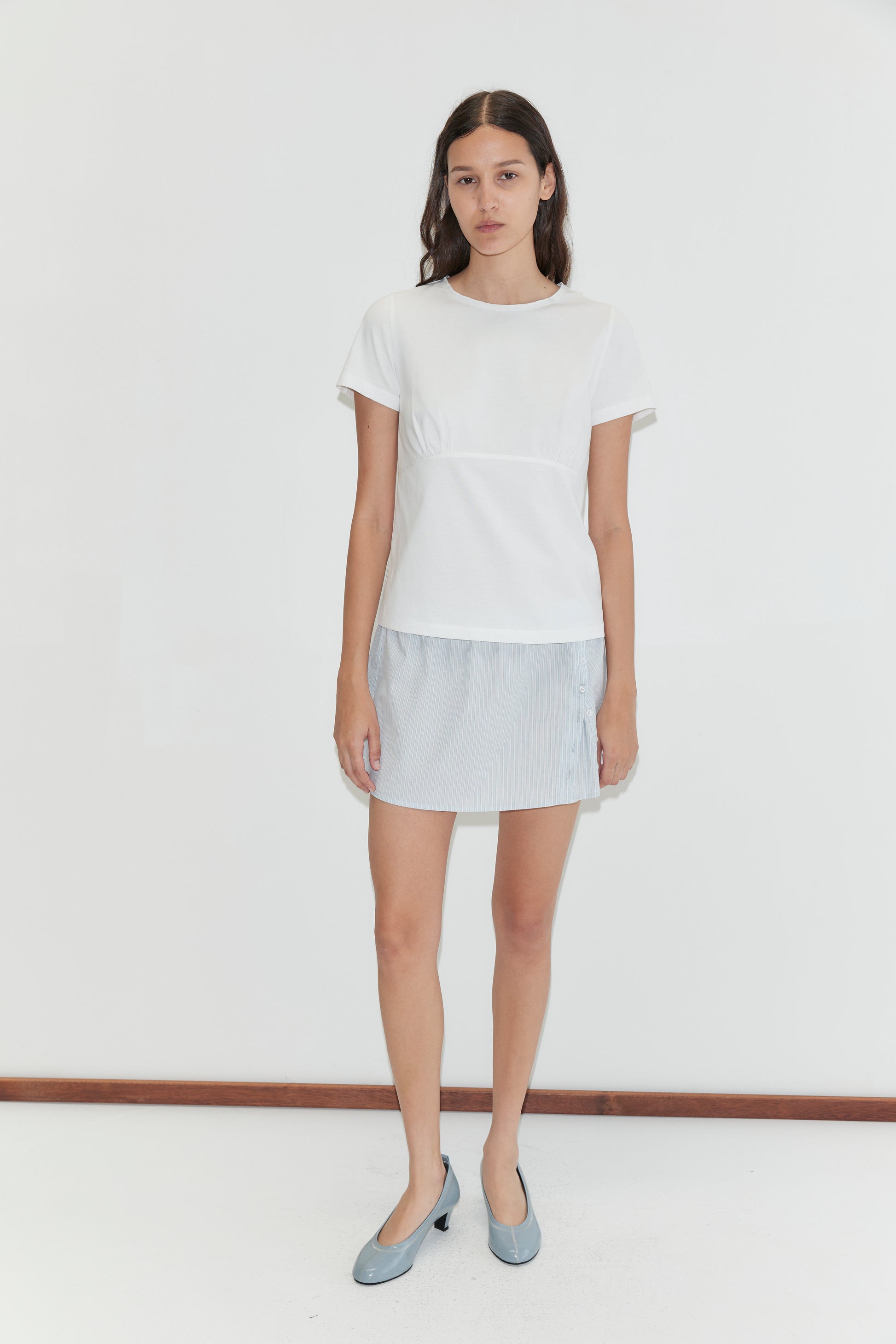 A woman with long dark hair and a neutral expression stands against a plain white background wearing the Pleat Split Skirt - Mist Stripe by Deiji Studios, a short-sleeved white top, and light blue flat shoes.