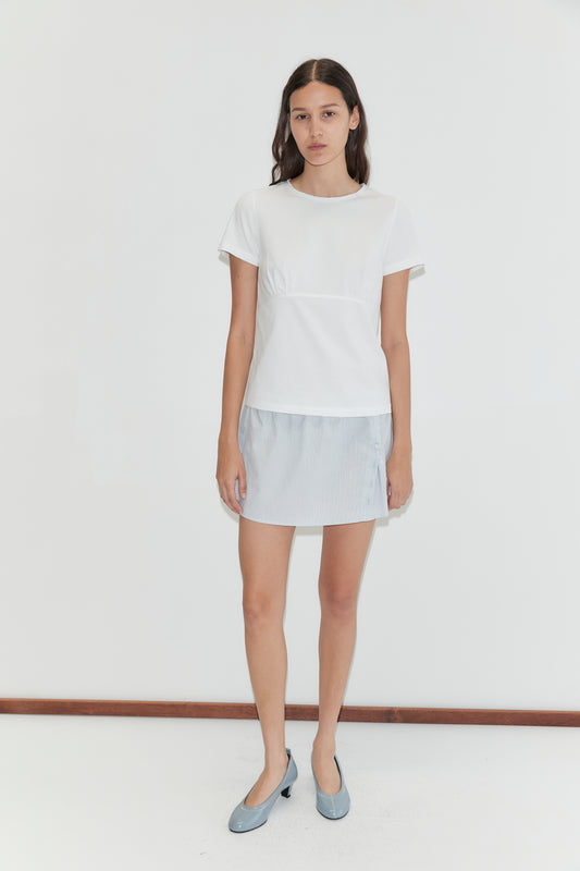 A woman with long dark hair and a neutral expression stands against a plain white background wearing the Pleat Split Skirt - Mist Stripe by Deiji Studios, a short-sleeved white top, and light blue flat shoes.