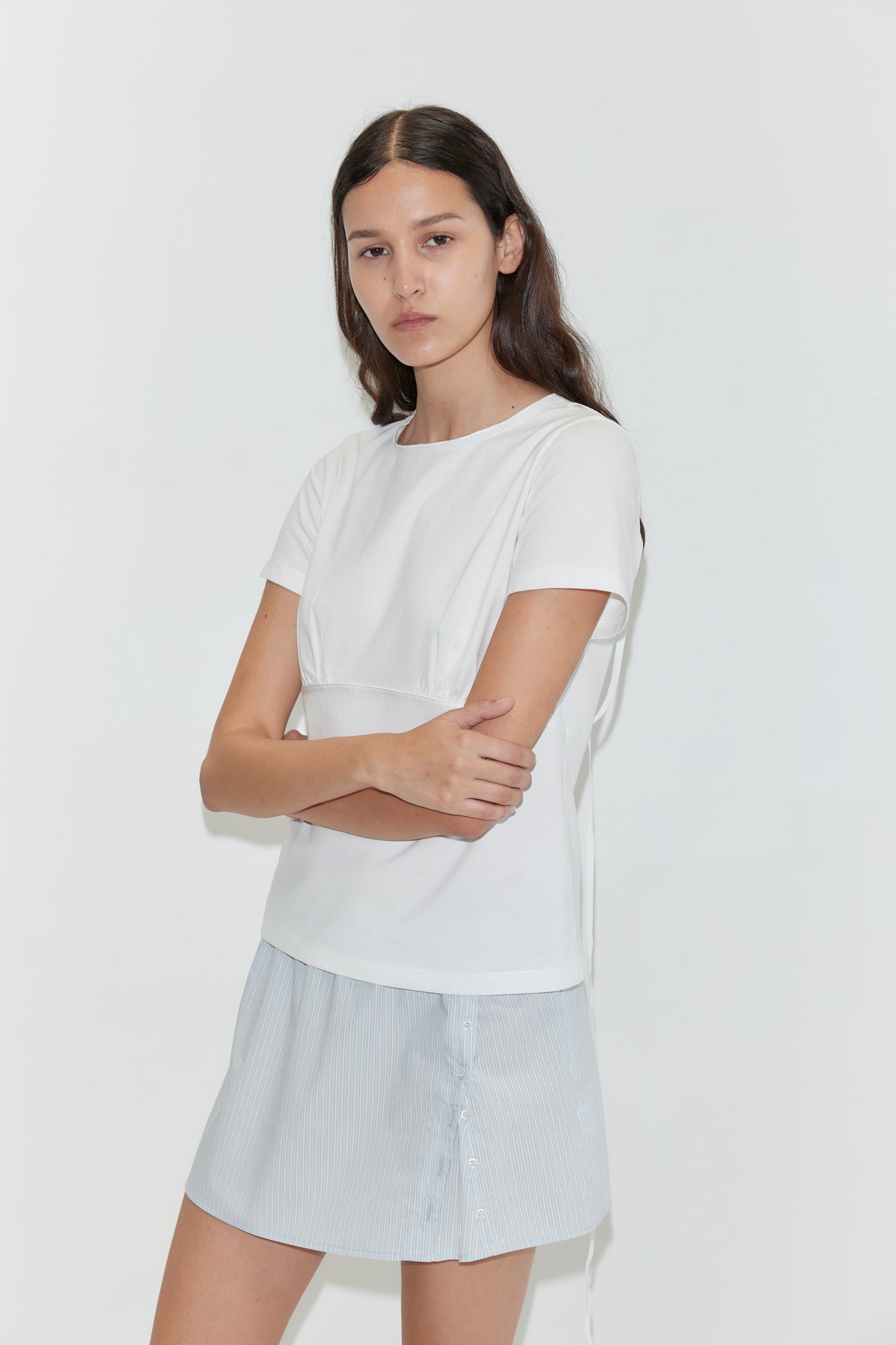 A young woman with long brown hair stands against a white background, arms crossed and neutral-faced, wearing a white short-sleeved top and the Pleat Split Skirt - Mist Stripe in light blue and white cotton poplin.