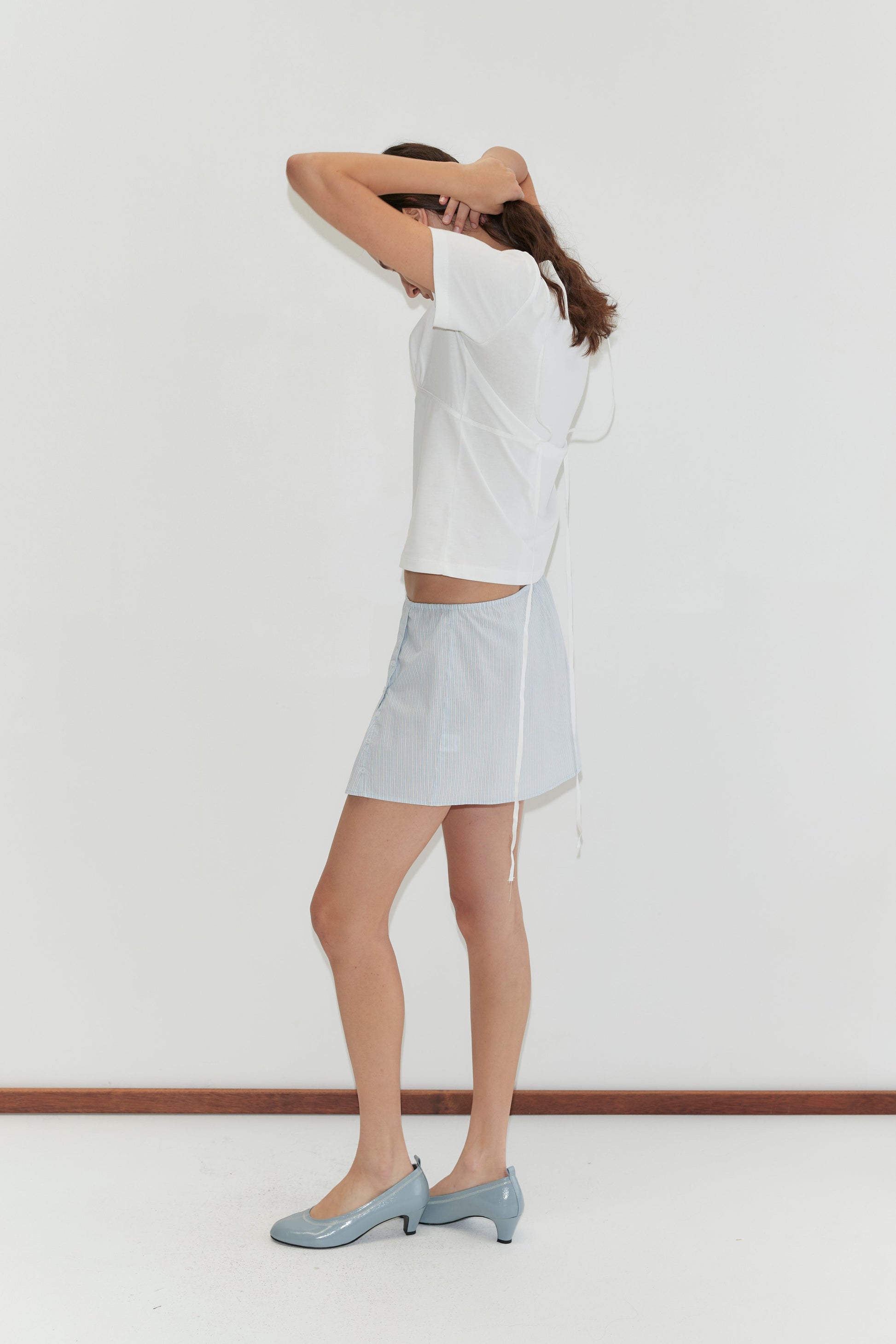 A woman poses against a white wall, wearing a white short-sleeve top, the Pleat Split Skirt - Mist Stripe in light blue, and matching blue heels. She raises one arm to adjust her hair, partially covering her face.