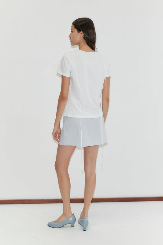 A woman faces away from the camera in a Tape Tee - White, a light blue mini skirt, and blue heels, set against a plain white background.