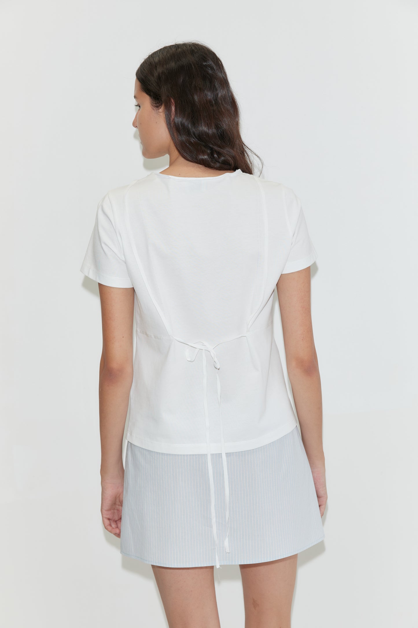 A woman with long, wavy dark hair faces away, wearing the Tape Tee - White by Deiji Studios and a light blue textured mini skirt, set against a plain white background.