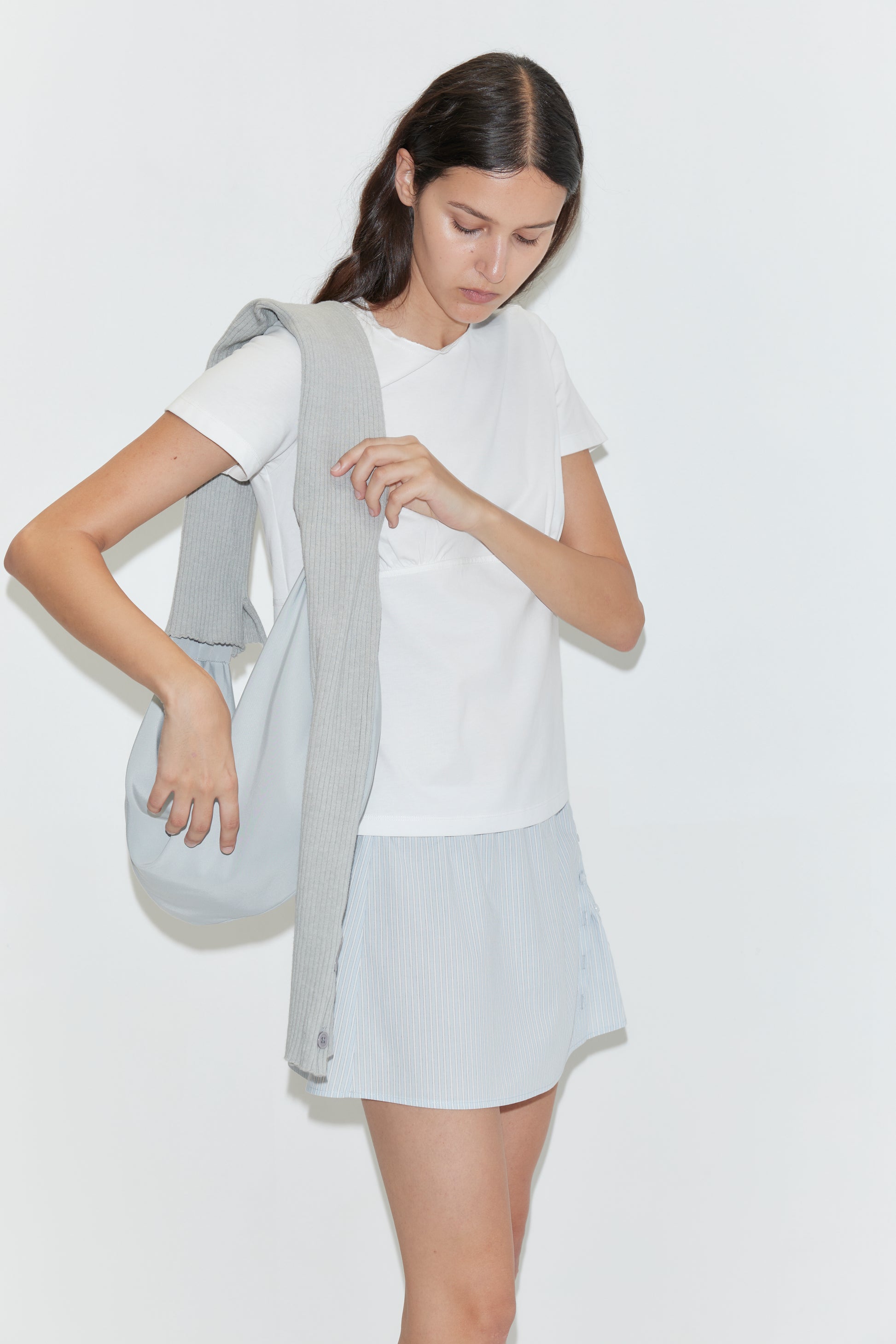 A woman with long dark hair stands against a plain white background, wearing a white t-shirt, baby blue mini skirt, gray sweater draped over her shoulder, and holding the Knit Strap Bag in Baby Blue.