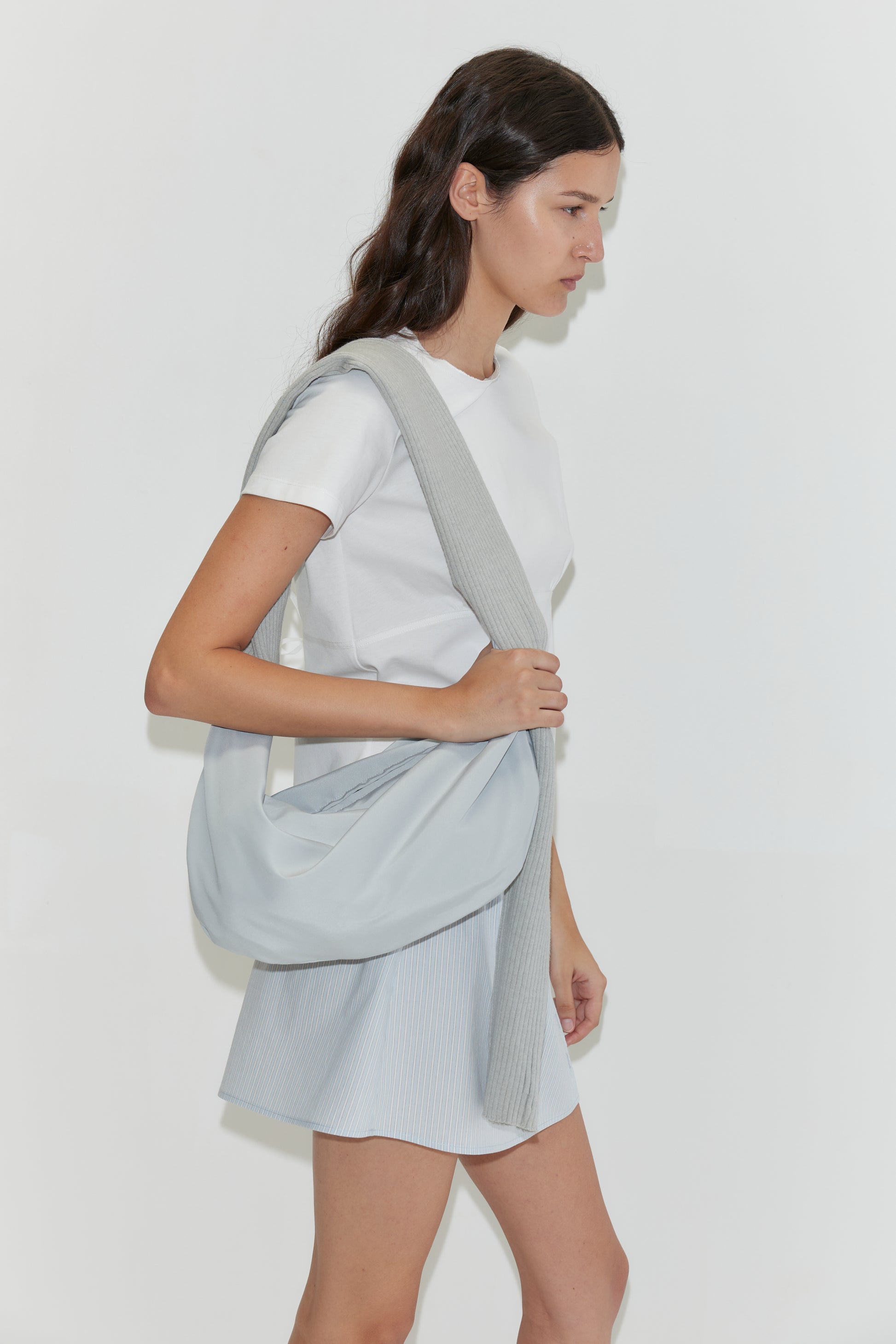 A woman with long dark hair walks against a white background, wearing a white t-shirt, a baby blue skirt, and carrying the Knit Strap Bag in Baby Blue.