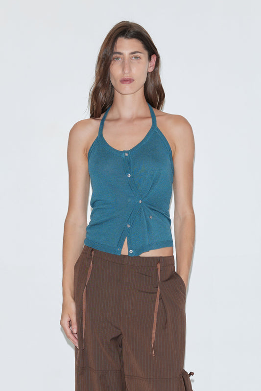 A woman wearing the Cross Top - Aqua, a fitted knit halter in sleeveless teal with a button-through front, paired with brown pinstripe pants featuring ribbon details, stands against a plain white background.