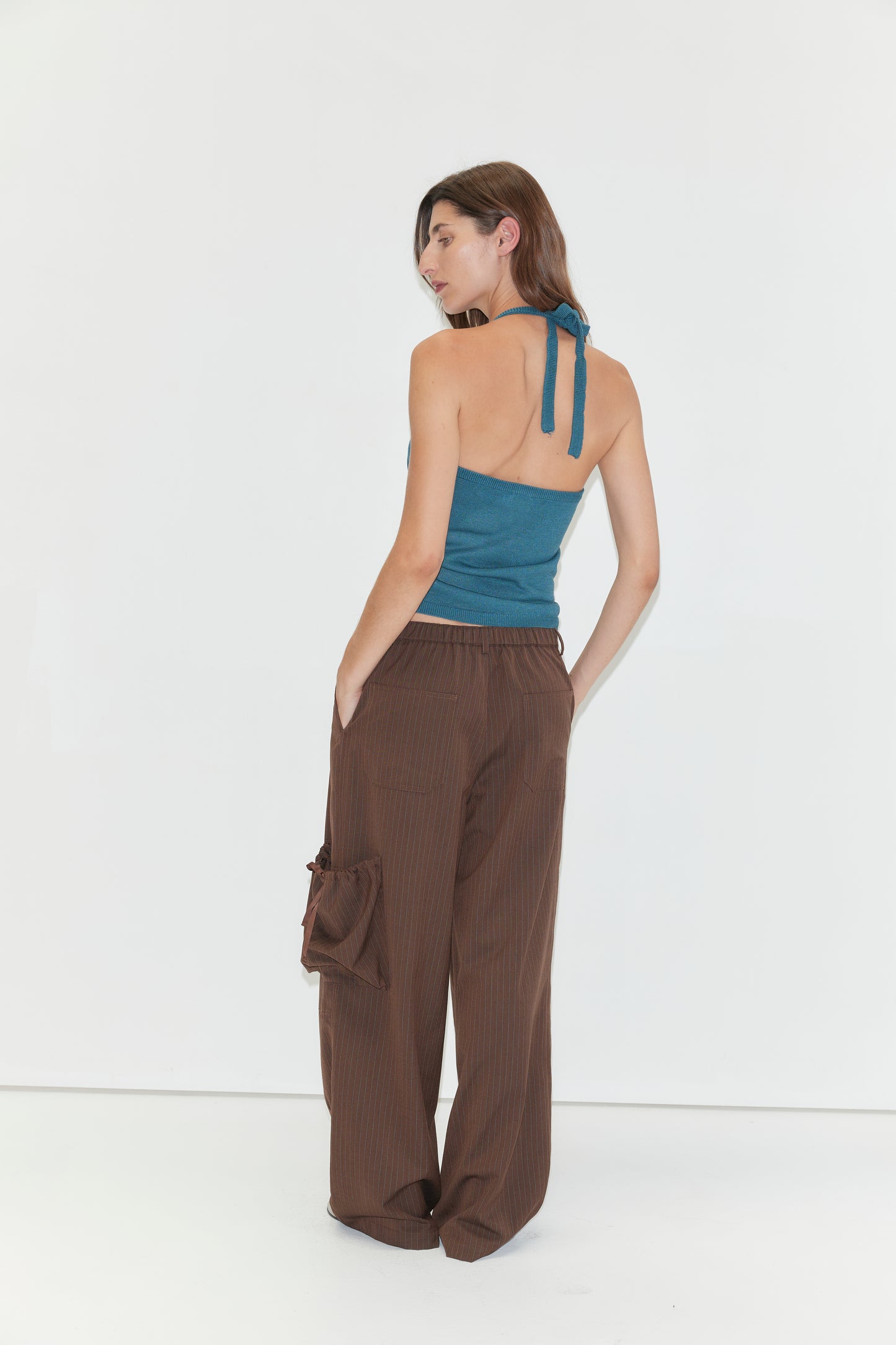 A woman with long brown hair poses in the fitted Cross Top - Aqua, a knit halter style featuring adjustable self-tie straps, paired with brown pinstripe cargo pants. With her hands in her pockets and set against a white backdrop, she glances over her shoulder to the left.