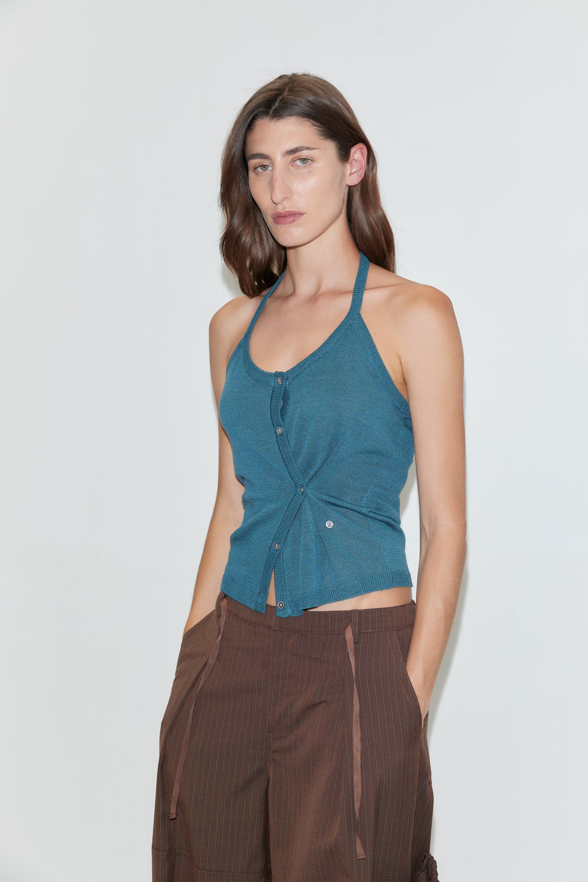 A woman with long brown hair wears the Cross Top in Aqua—a fitted knit halter top with adjustable self-tie straps—paired with brown pants, standing against a plain white background and looking at the camera with a neutral expression.