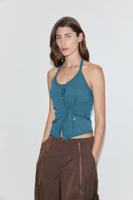 A woman with long brown hair wears the Cross Top in Aqua—a fitted knit halter top with adjustable self-tie straps—paired with brown pants, standing against a plain white background and looking at the camera with a neutral expression.