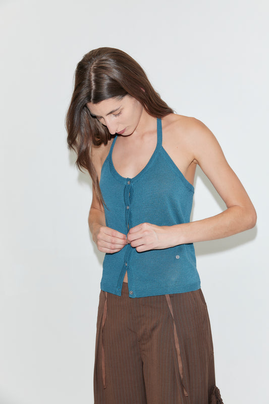 A woman with long brown hair looks down while buttoning the front of the Cross Top in Aqua, a blue fitted knit halter top with adjustable self-tie straps. She is wearing brown pants and standing against a plain white background.