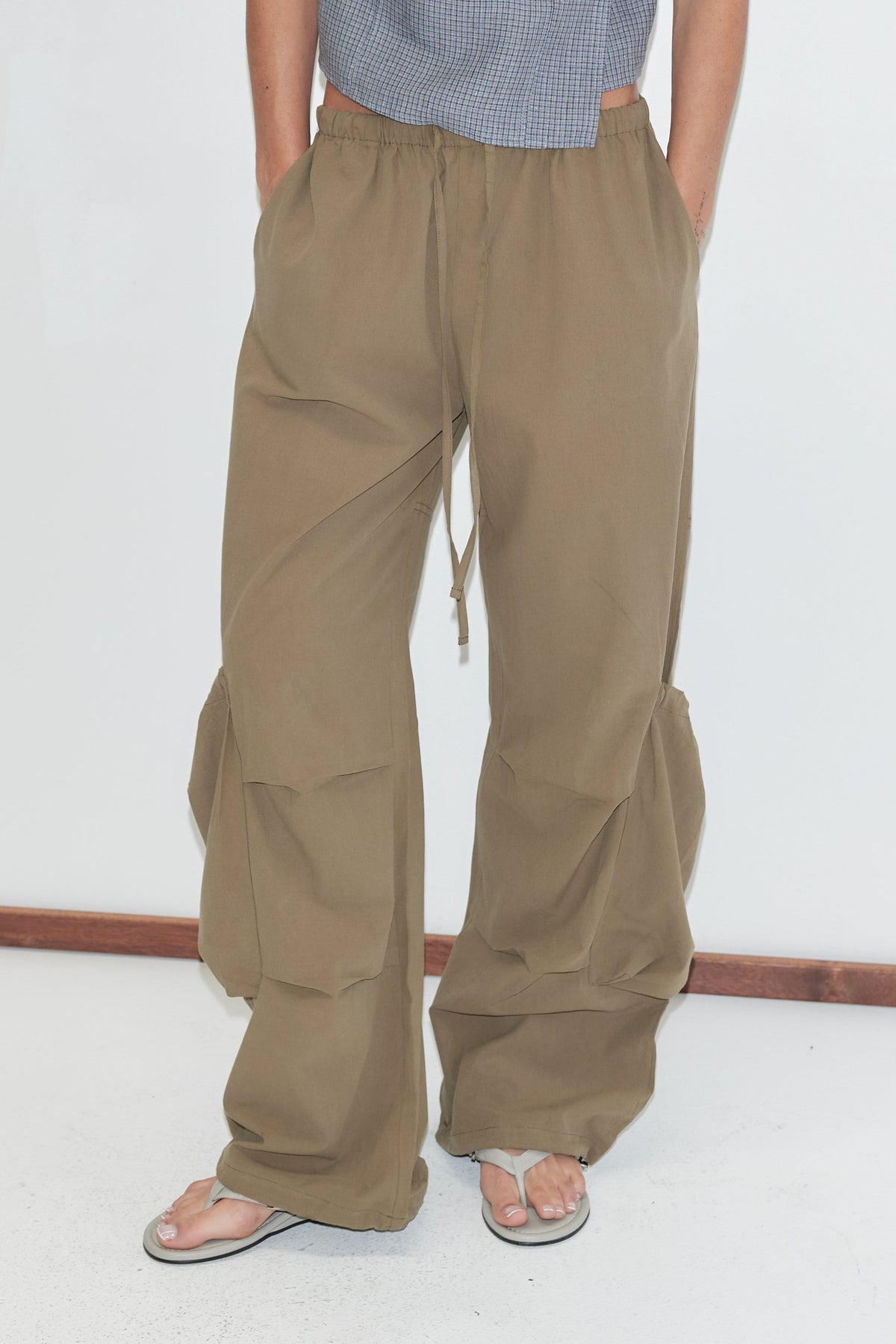A cropped view of the Drawcord Pocket Pant - Khaki by Deiji Studios, khaki wide-leg trousers with a drawcord waist, side pockets and drawcord ties gathering the lower legs, worn with flip-flops against a plain white background.