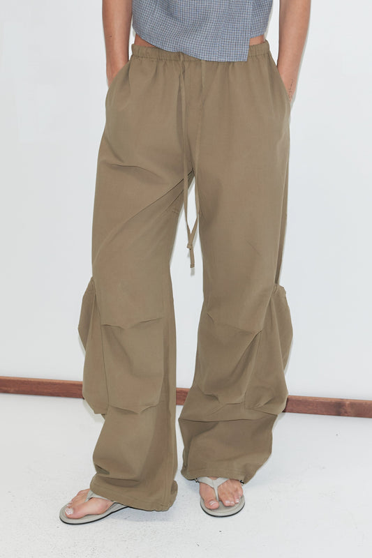 A cropped view of the Drawcord Pocket Pant - Khaki by Deiji Studios, khaki wide-leg trousers with a drawcord waist, side pockets and drawcord ties gathering the lower legs, worn with flip-flops against a plain white background.