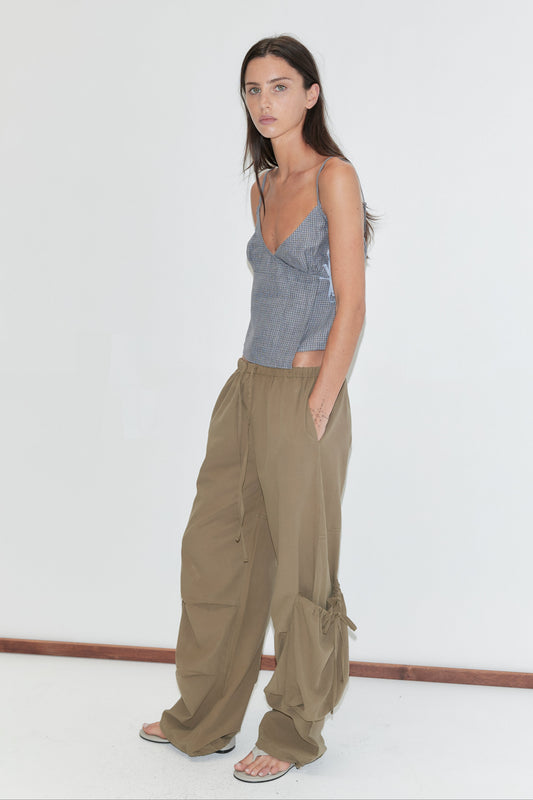 A woman with long dark hair stands in profile against a plain white background wearing the Drawcord Pocket Pant - Khaki by Deiji Studios, khaki wide-leg trousers, with a blue check cami top and flip-flops.