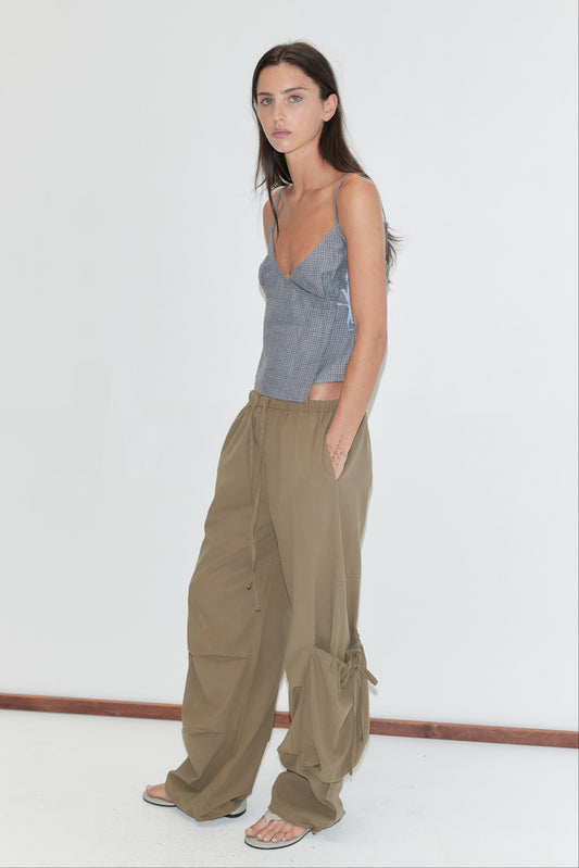 A woman with long dark hair stands in profile against a plain white background wearing the Wrapping Top - Indigo Check by Deiji Studios, an indigo micro-check linen cami, with khaki drawcord trousers and flip-flops.