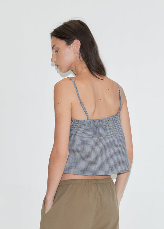 A woman with long dark hair stands with her back to the camera against a plain white background wearing the Wrapping Top - Indigo Check by Deiji Studios, an indigo micro-check linen cami with a gathered back, with khaki trousers.