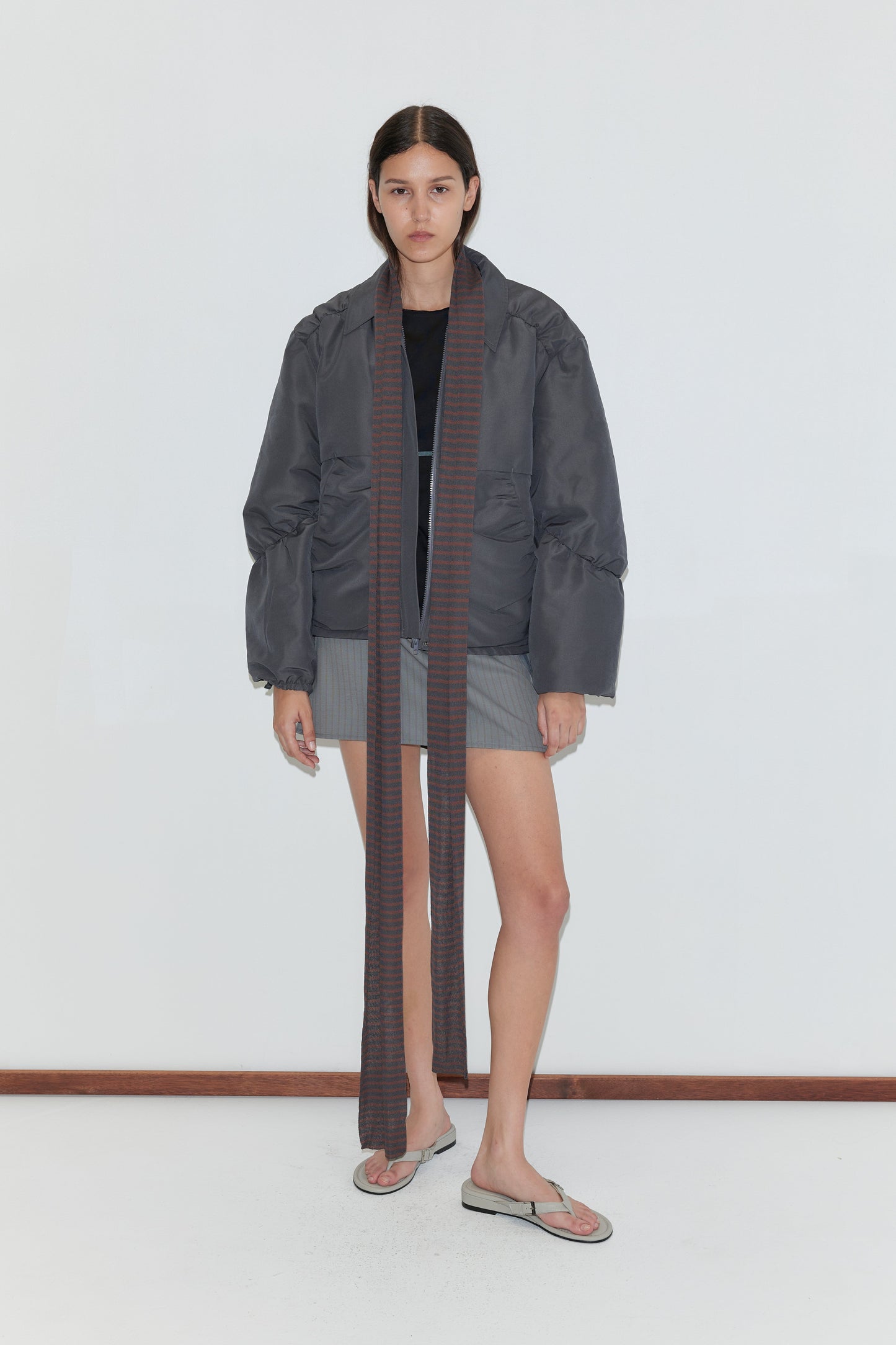 A woman with straight dark hair stands against a white wall, wearing a gray oversized puffer jacket, short gray skirt, black top, flip-flops, and the Knit Scarf - Brown Grey Stripe. She has a neutral expression.