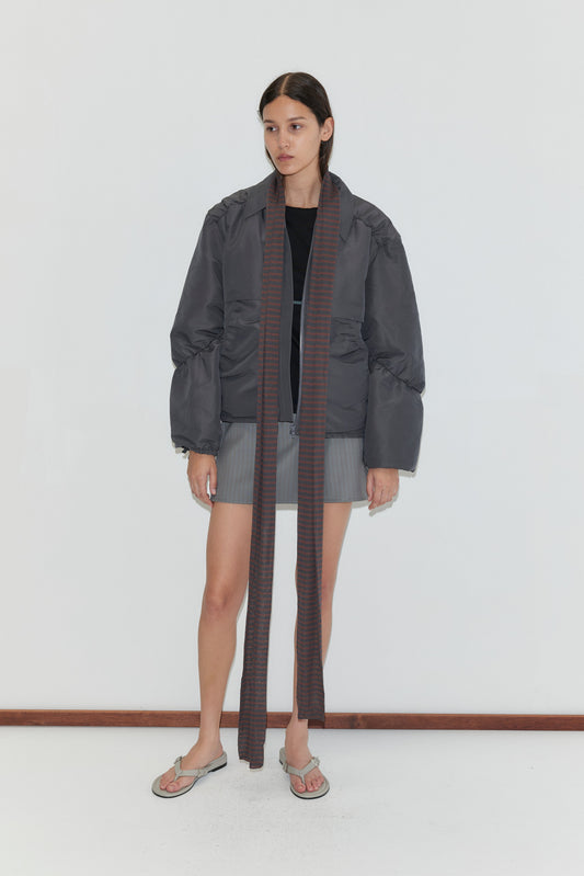 A woman with straight brown hair and a neutral expression stands against a white background, wearing a gray puffer jacket, black top, gray shorts, flip-flops, and the Knit Scarf - Brown Grey Stripe.