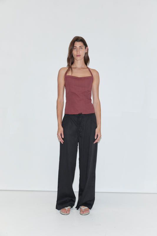 A person stands against a plain white background wearing a sleeveless maroon top, loose wide-leg Tencel Pant in Black by Deiji Studios, and sandals. They have long brown hair and a neutral expression, with arms relaxed at their sides.