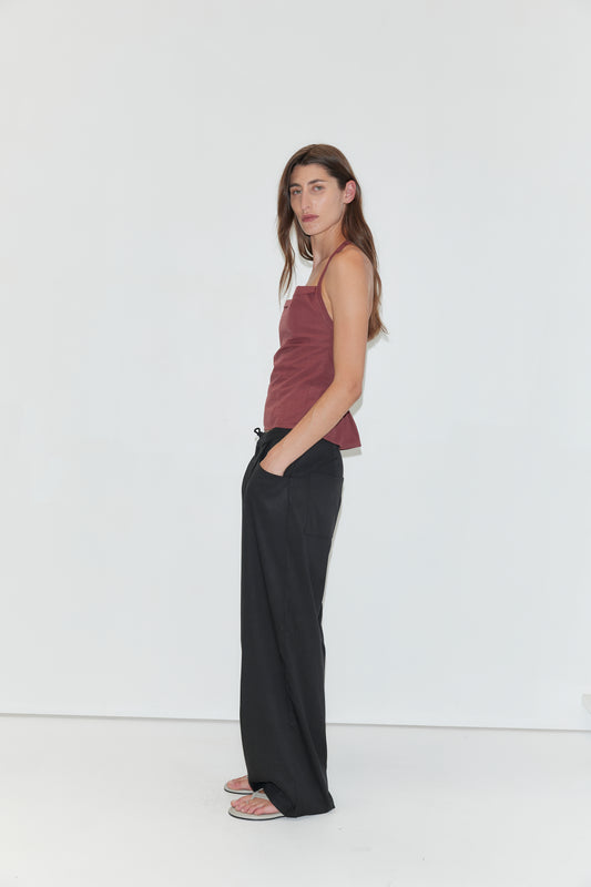 A woman with long brown hair stands in profile against a plain white background. She is wearing a sleeveless ribbed dark red top paired with the Tencel Pant in Black—loose-fitting, wide-leg trousers—with her hand tucked casually in her pocket. Simple sandals complete the outfit, creating a relaxed yet refined look reminiscent of Deiji Studios’ effortless style.