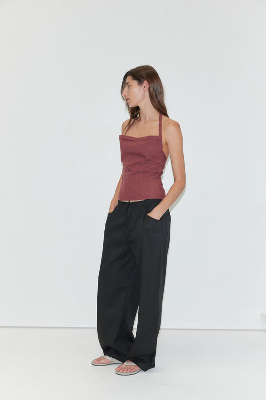 A woman stands against a white background, wearing the Folded Top in Rust Stripe, paired with loose black pants and sandals. She has long brown hair and looks to her left with her hands in her pockets.