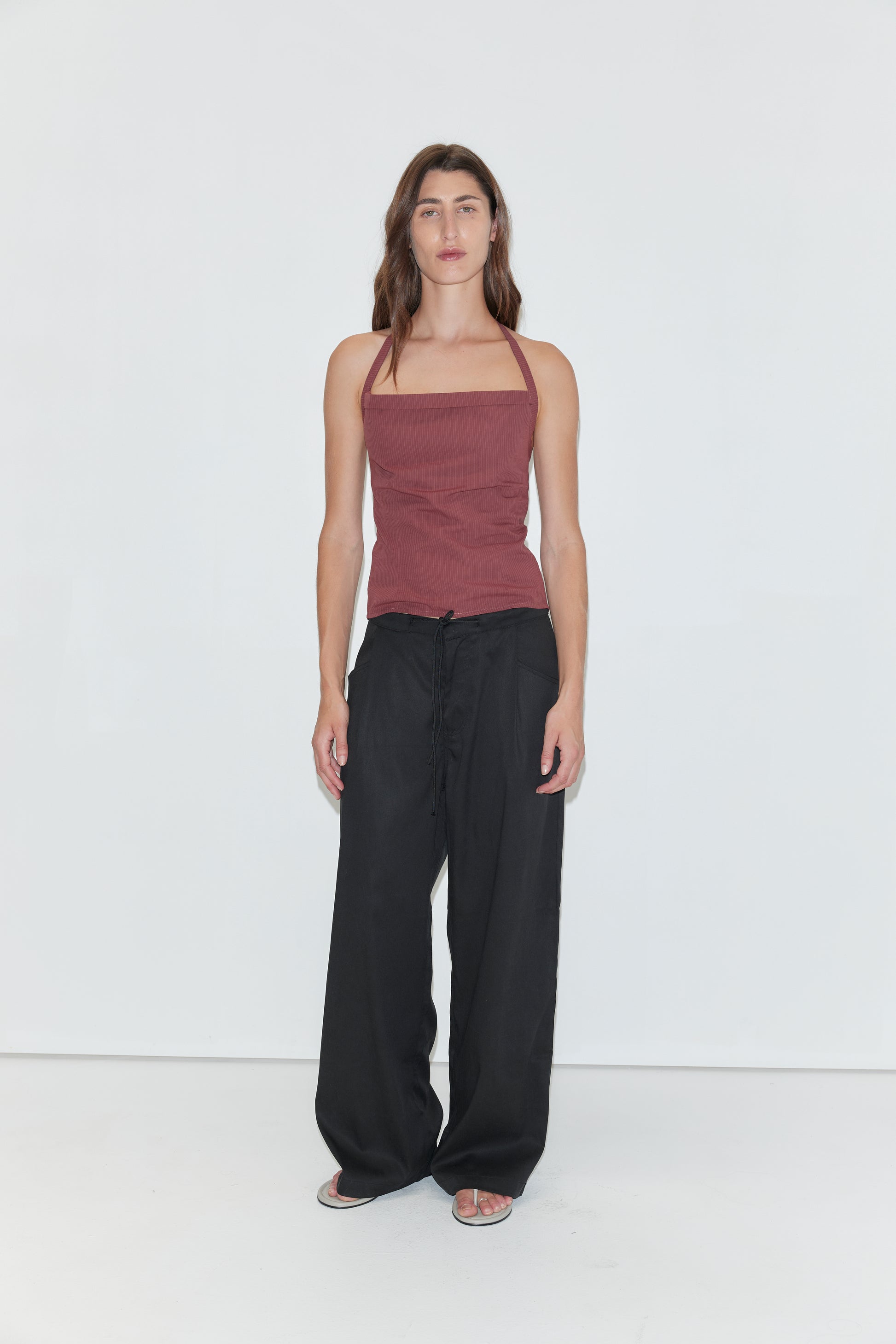 A person with long brown hair stands against a plain white background, wearing the Folded Top in Rust Stripe by Deiji Studios, paired with loose black pants and sandals.