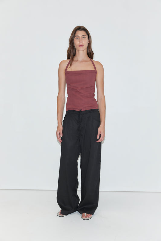 A person with long brown hair stands against a plain white background, wearing the Folded Top in Rust Stripe by Deiji Studios, paired with loose black pants and sandals.