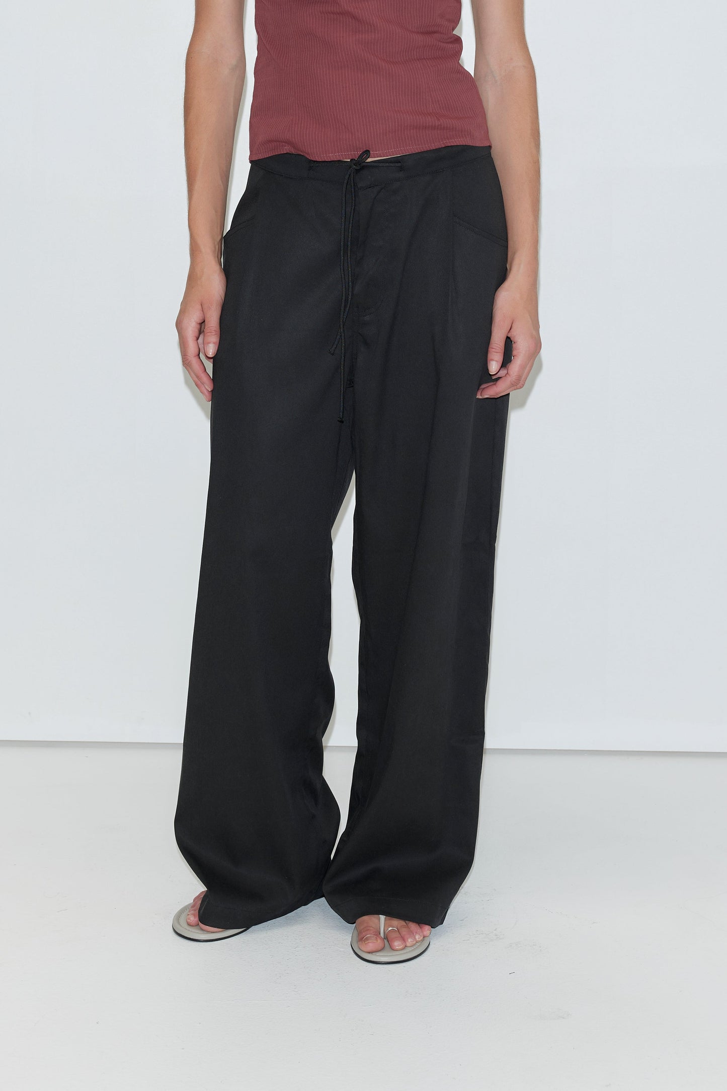 A person wearing a sleeveless maroon top, loose black Tencel Pant - Black from Deiji Studios, and white flip-flops stands against a plain white background.