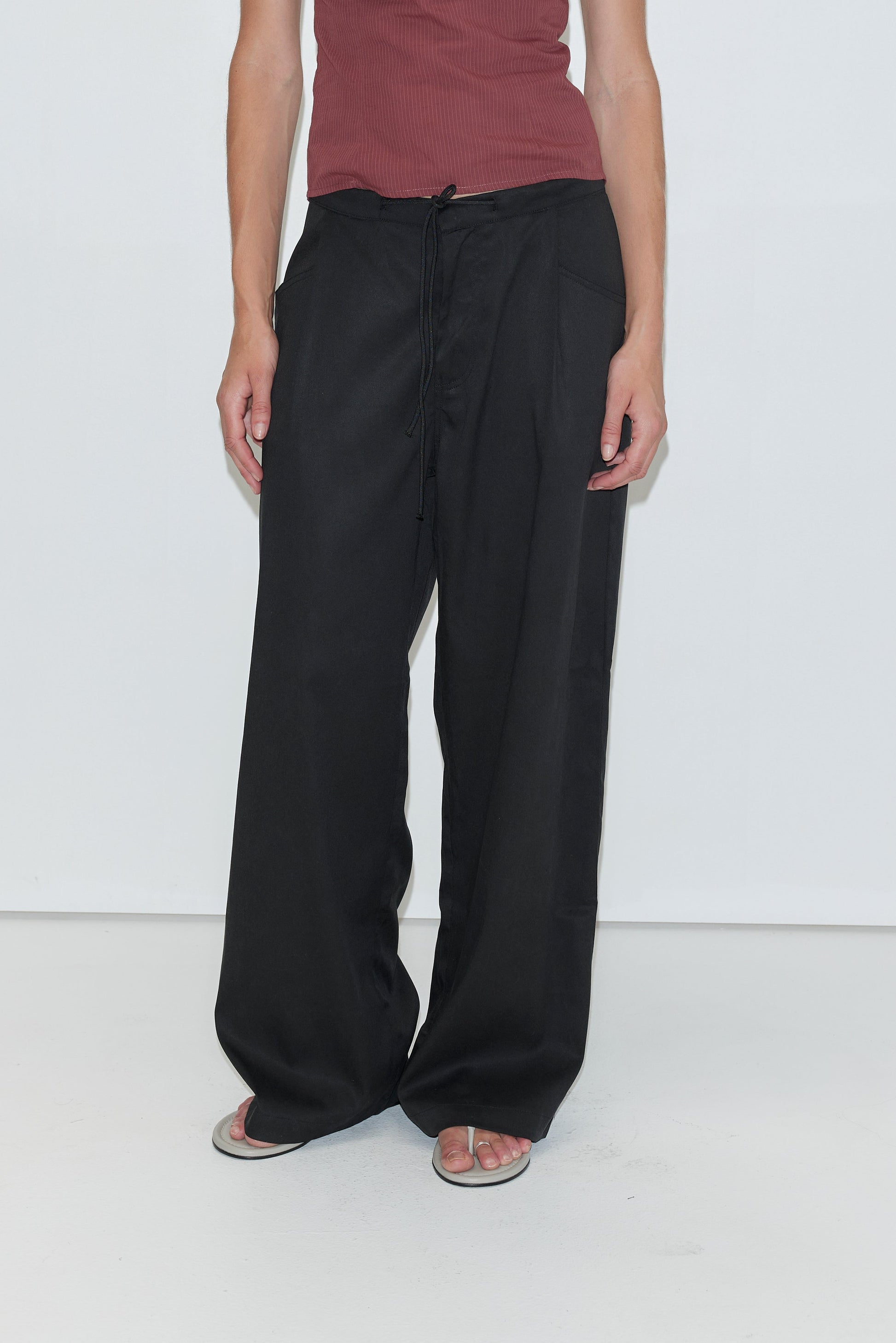 A person wearing a sleeveless maroon top, loose black Tencel Pant - Black from Deiji Studios, and white flip-flops stands against a plain white background.