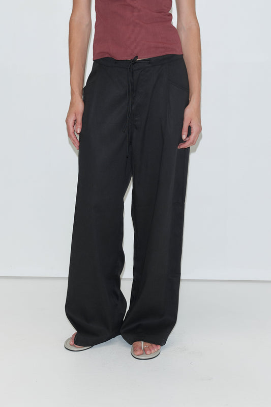 A person wearing a sleeveless maroon top, loose black Tencel Pant - Black from Deiji Studios, and white flip-flops stands against a plain white background.