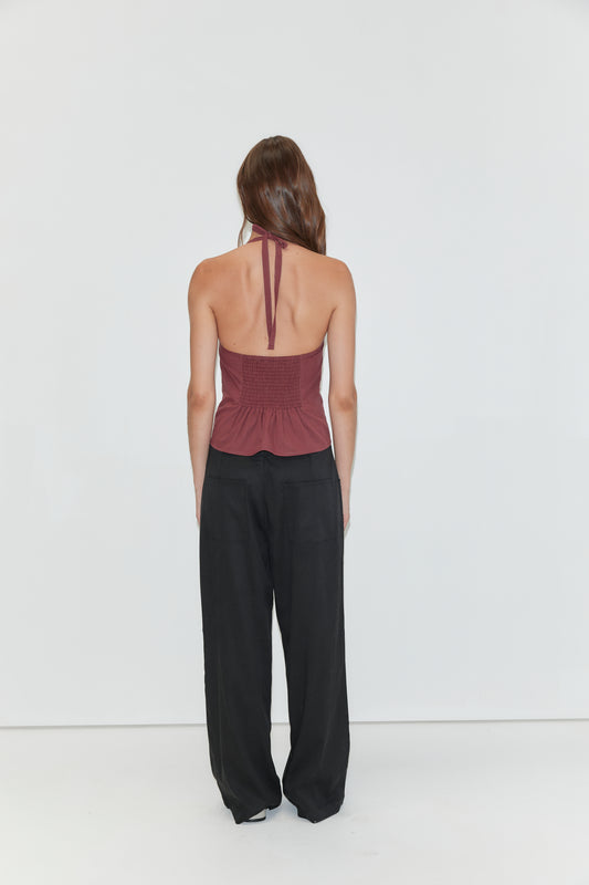 A woman stands facing away, wearing a maroon halter top with an open back and loose black Tencel Pant by Deiji Studios, against a plain white background.