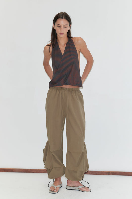 A woman with long dark hair stands against a plain white background wearing the Drawcord Pocket Pant - Khaki by Deiji Studios, khaki wide-leg trousers with drawcord-gathered lower legs, with a brown striped halter top and grey flip-flops.