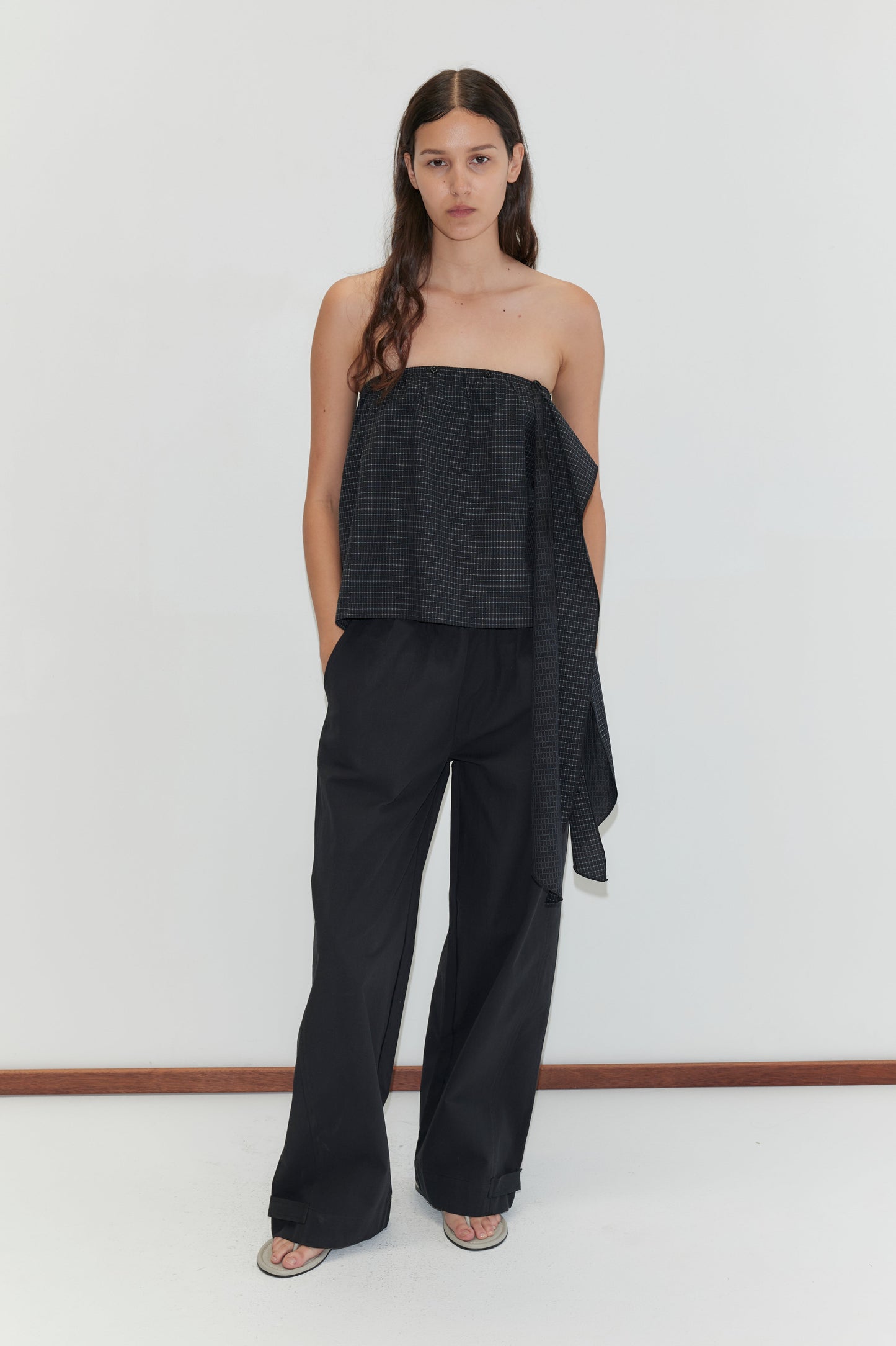 A woman with long brown hair wears Deiji Studios' Flow Top - Mono Grid, a strapless cotton top with a dark grid pattern and asymmetrical drape, paired with wide-leg black pants and sandals. She stands against a plain white background, hands in her pockets.