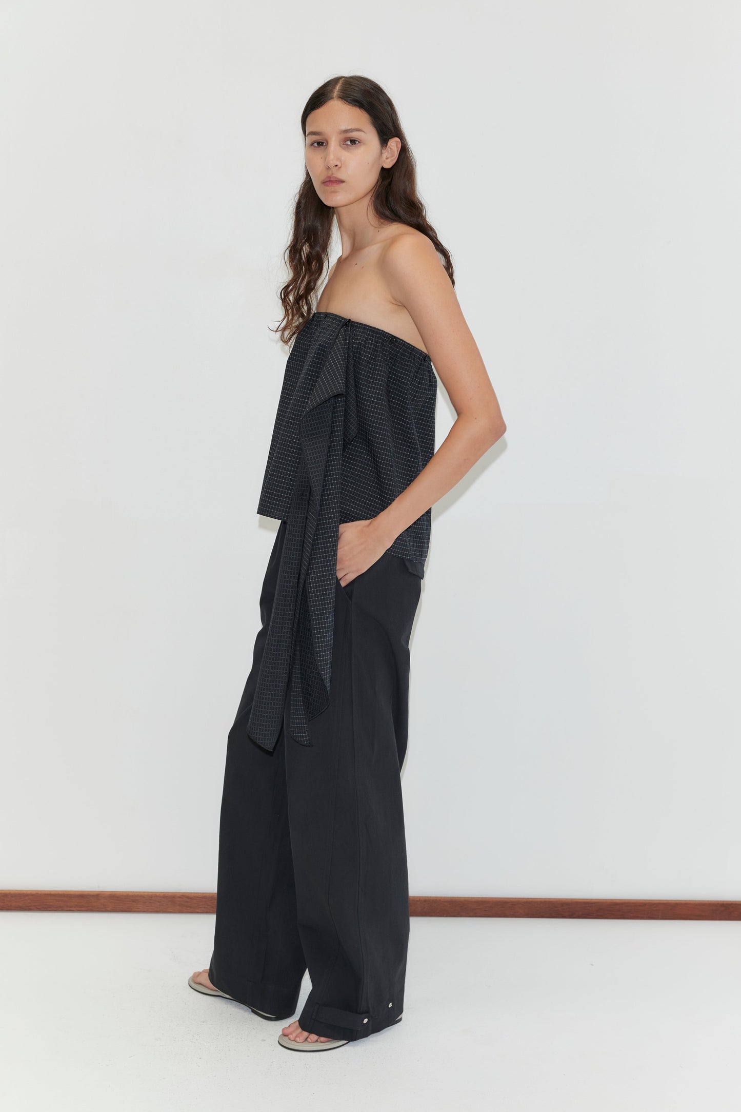 A person with long, wavy hair stands sideways against a plain white wall, wearing the Flow Top - Mono Grid by Deiji Studios with a bow detail and wide-leg black pants. They complete the look with white open-toe shoes.