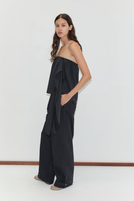 A person with long, wavy hair stands sideways against a plain white wall, wearing the Flow Top - Mono Grid by Deiji Studios with a bow detail and wide-leg black pants. They complete the look with white open-toe shoes.