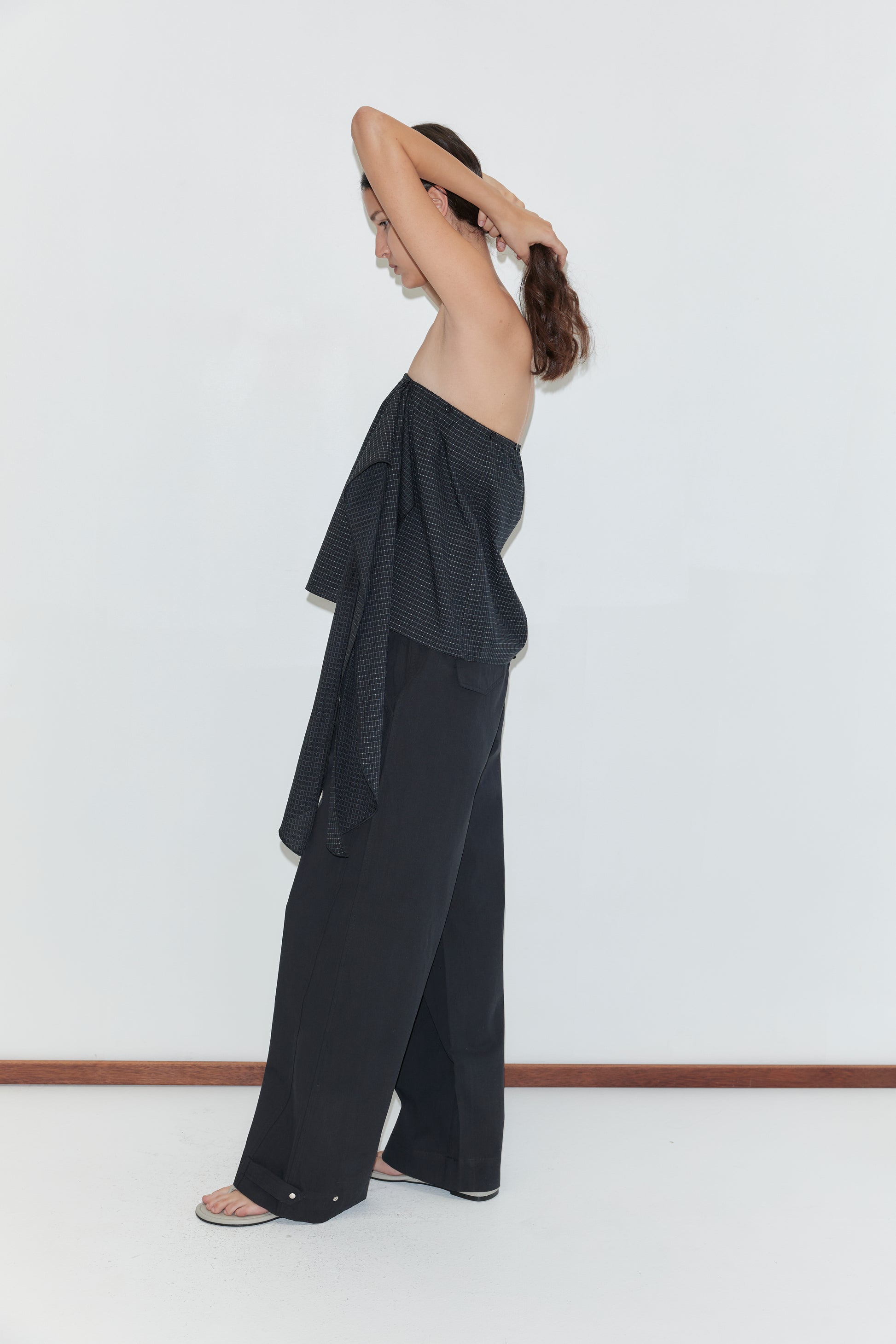 A person stands sideways against a white wall, wearing a loose black strapless top, Snap Tab Pant - Black by Deiji Studios, and sandals, with their arms raised to tie their hair back.