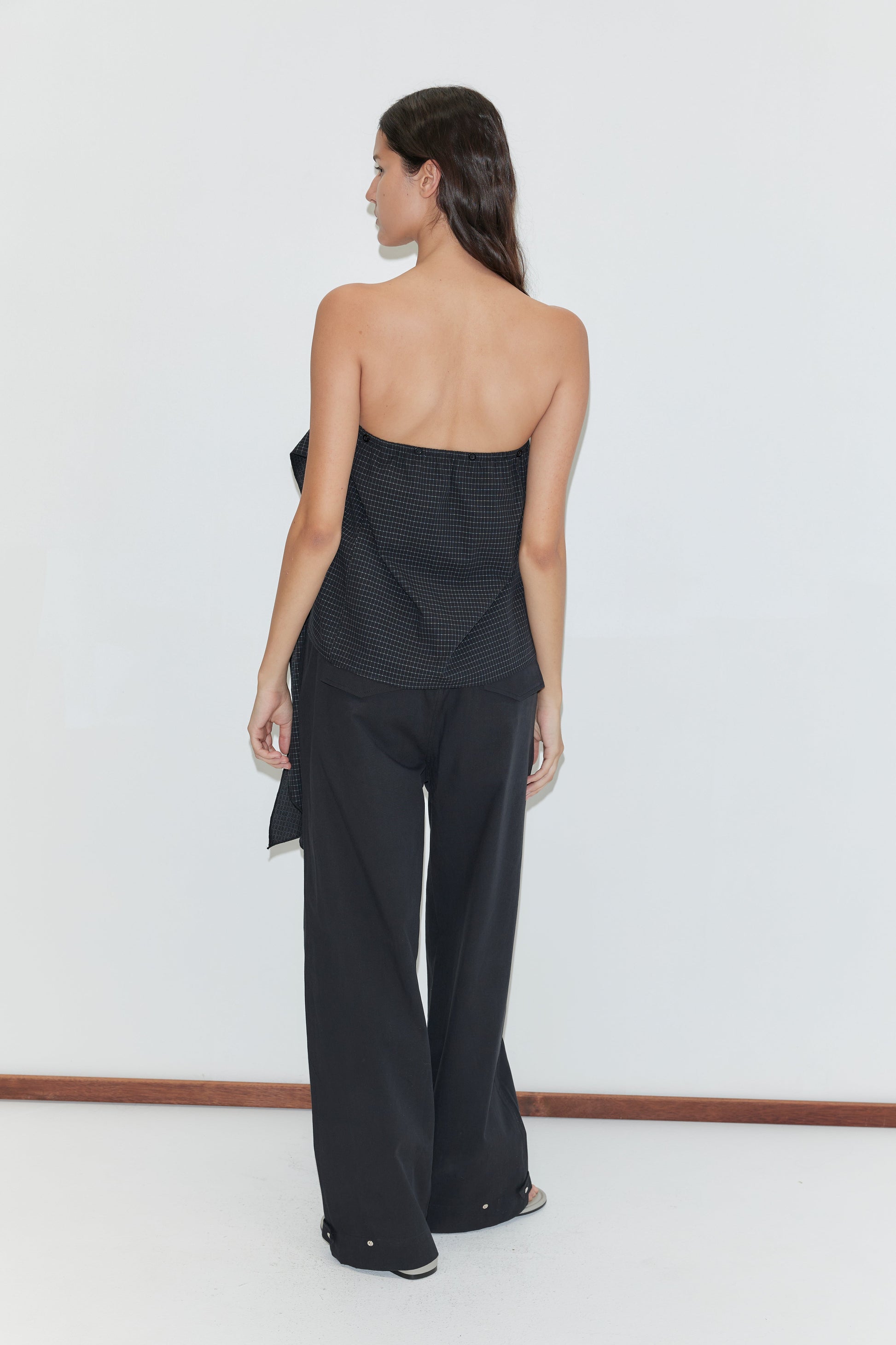 A woman with long dark hair stands against a white background, wearing Deiji Studios' Flow Top - Mono Grid—a strapless cotton top with a textured pattern—paired with loose black pants.