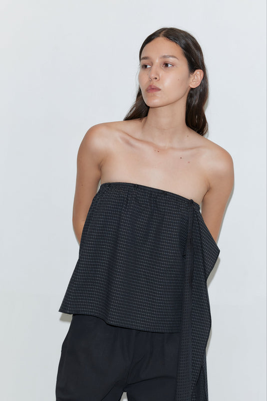 A woman with long dark hair stands against a plain light background, wearing the Flow Top - Mono Grid by Deiji Studios and loose black pants, looking slightly to her right with a neutral expression.