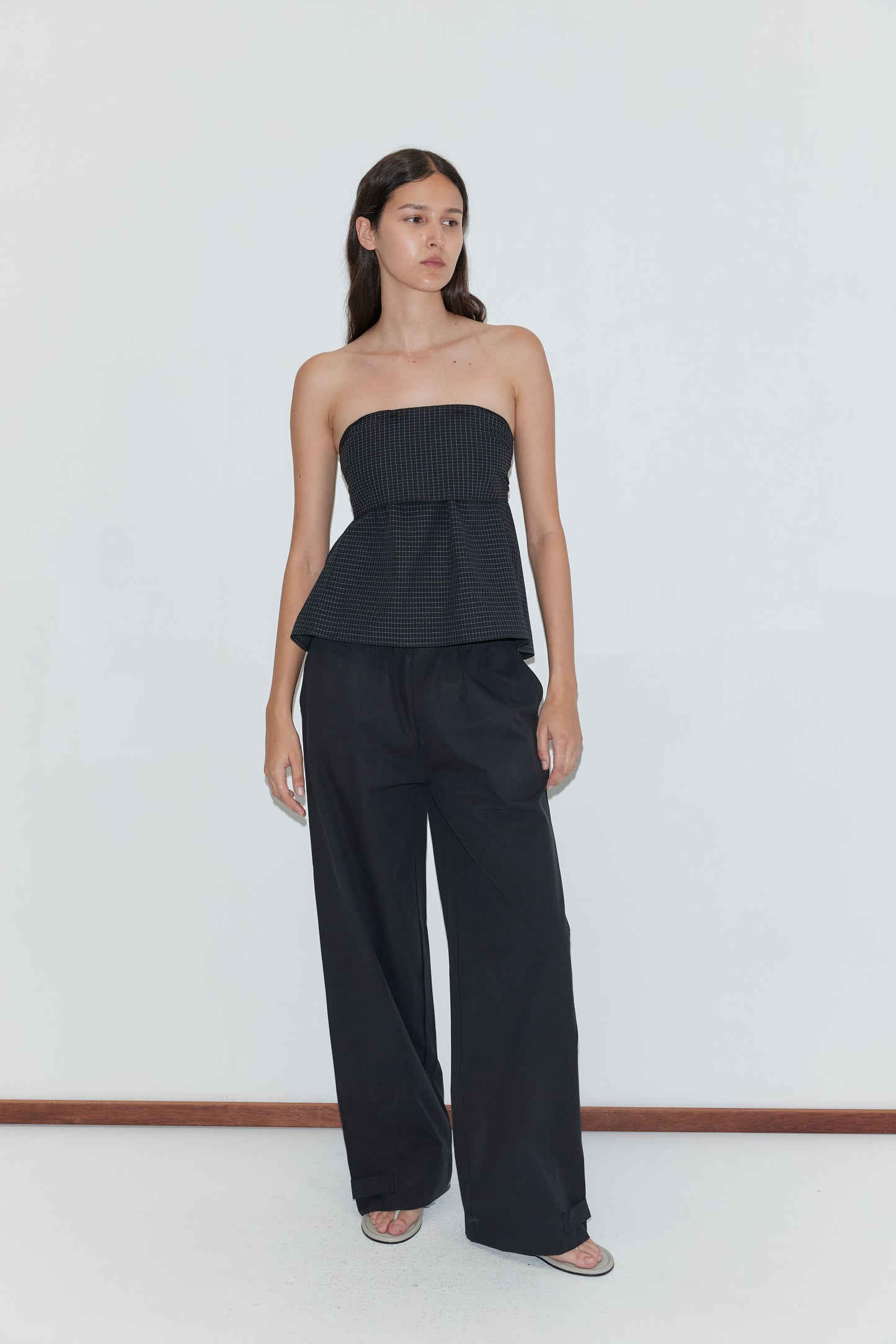 A woman stands against a white wall wearing a black strapless, textured peplum top and Snap Tab Pant - Black by Deiji Studios, with long brown hair down and open-toe sandals.