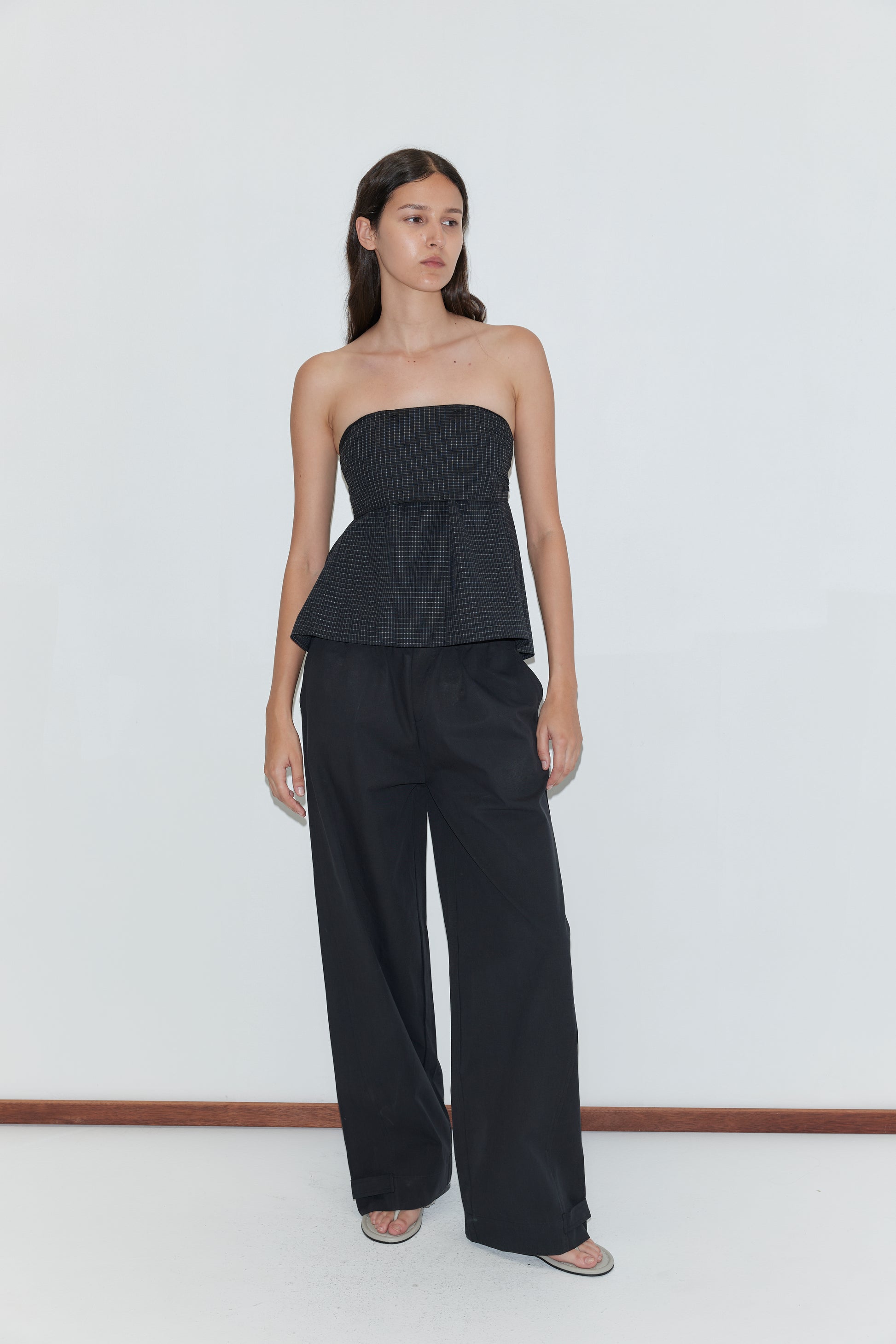 A woman stands against a white wall wearing a black strapless, textured peplum top and Snap Tab Pant - Black by Deiji Studios, with long brown hair down and open-toe sandals.