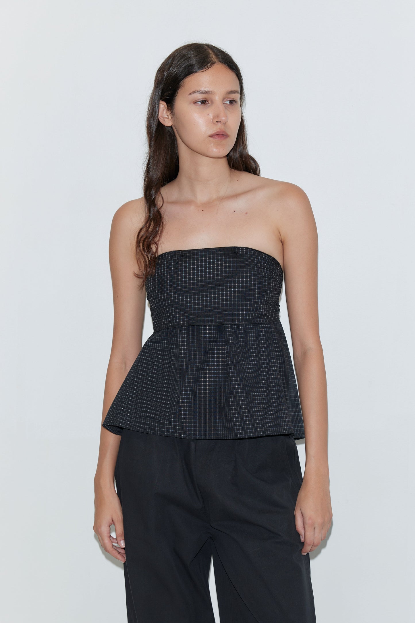 A woman with long brown hair wears the Flow Top - Mono Grid by Deiji Studios, styled with loose black pants and standing against a plain light background.