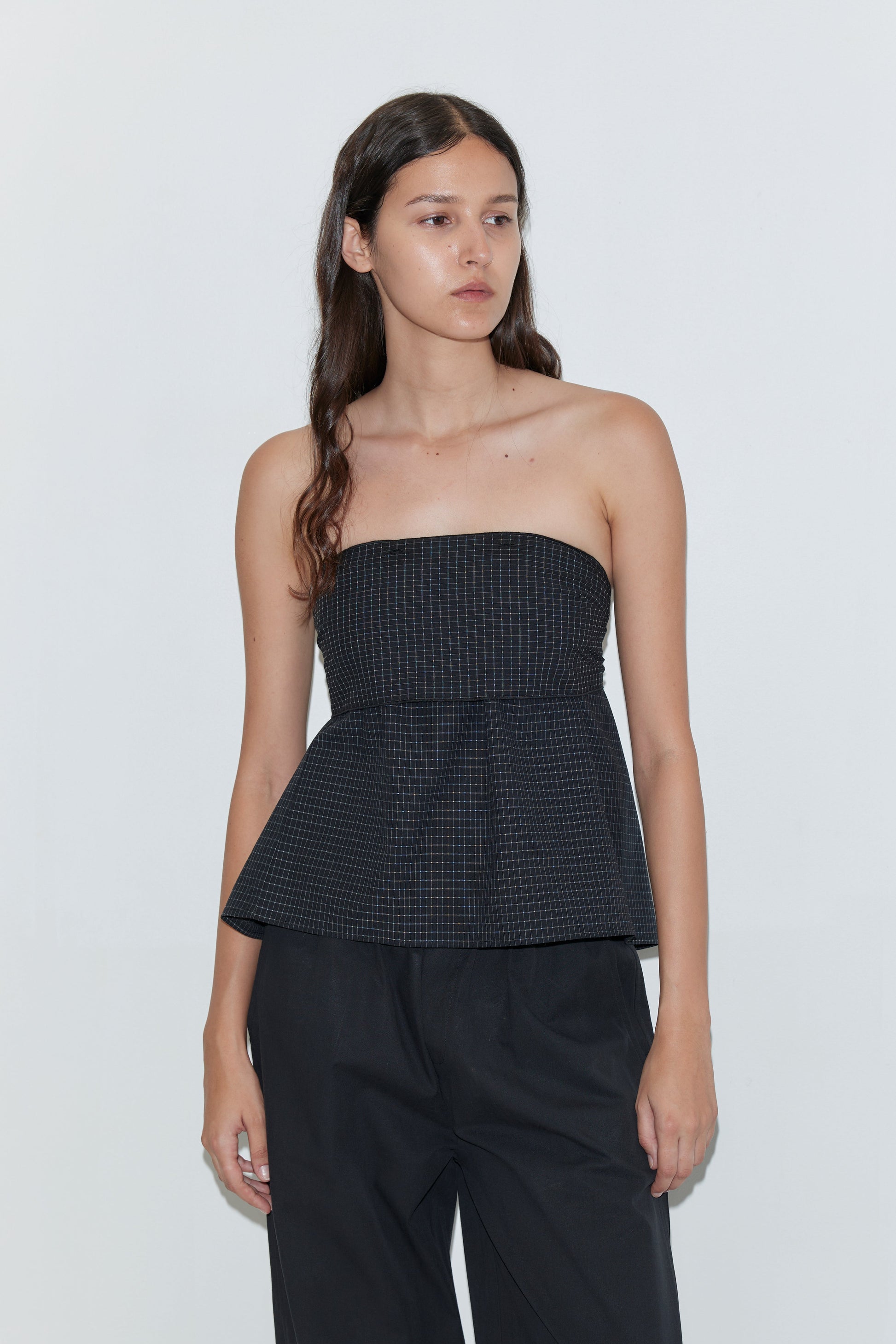 A woman with long brown hair wears the Flow Top - Mono Grid by Deiji Studios, styled with loose black pants and standing against a plain light background.